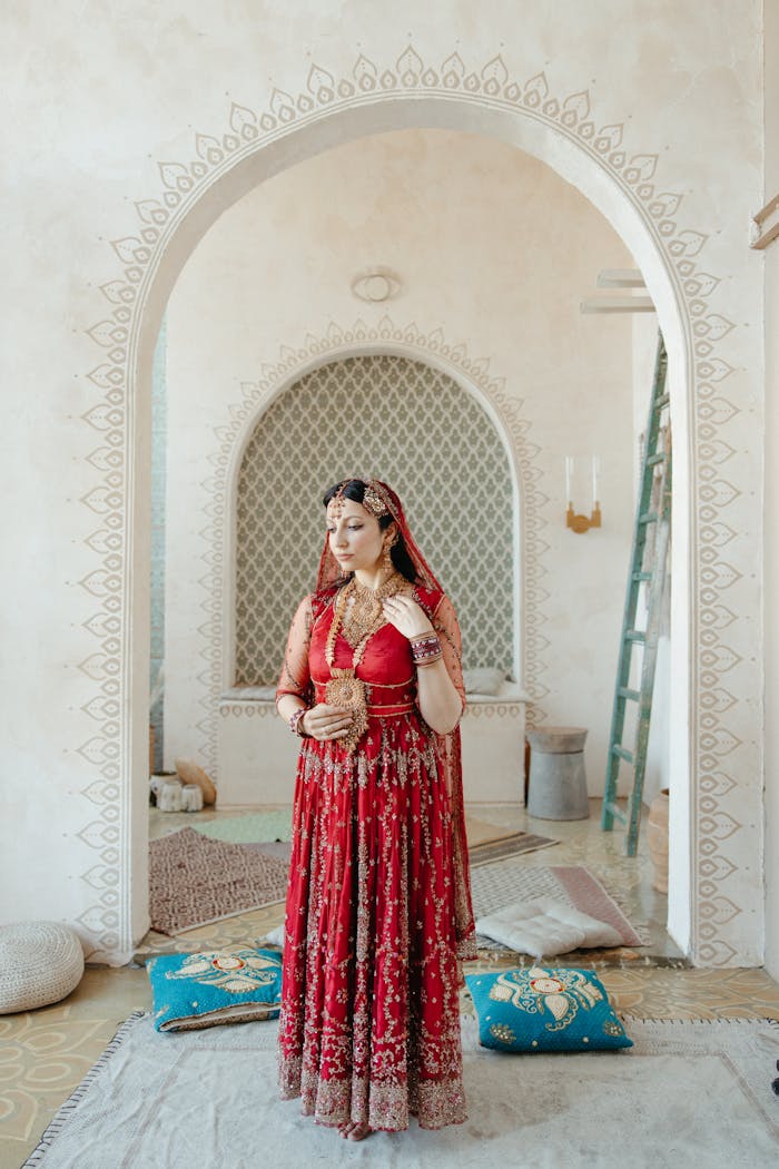 Indian woman in a red sari standing indoors, adorned with jewelry, showcasing cultural heritage.